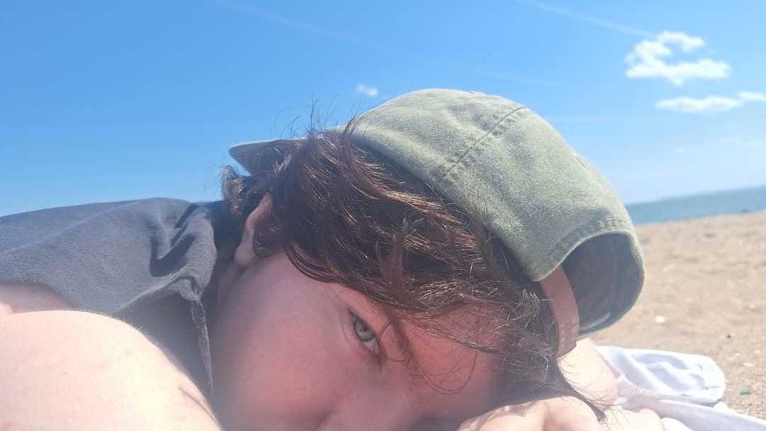 a picture of rhys, a boy with fair skin and brown hair tucked under a green baseball cap, from the shoulders up, laying on a beach.
